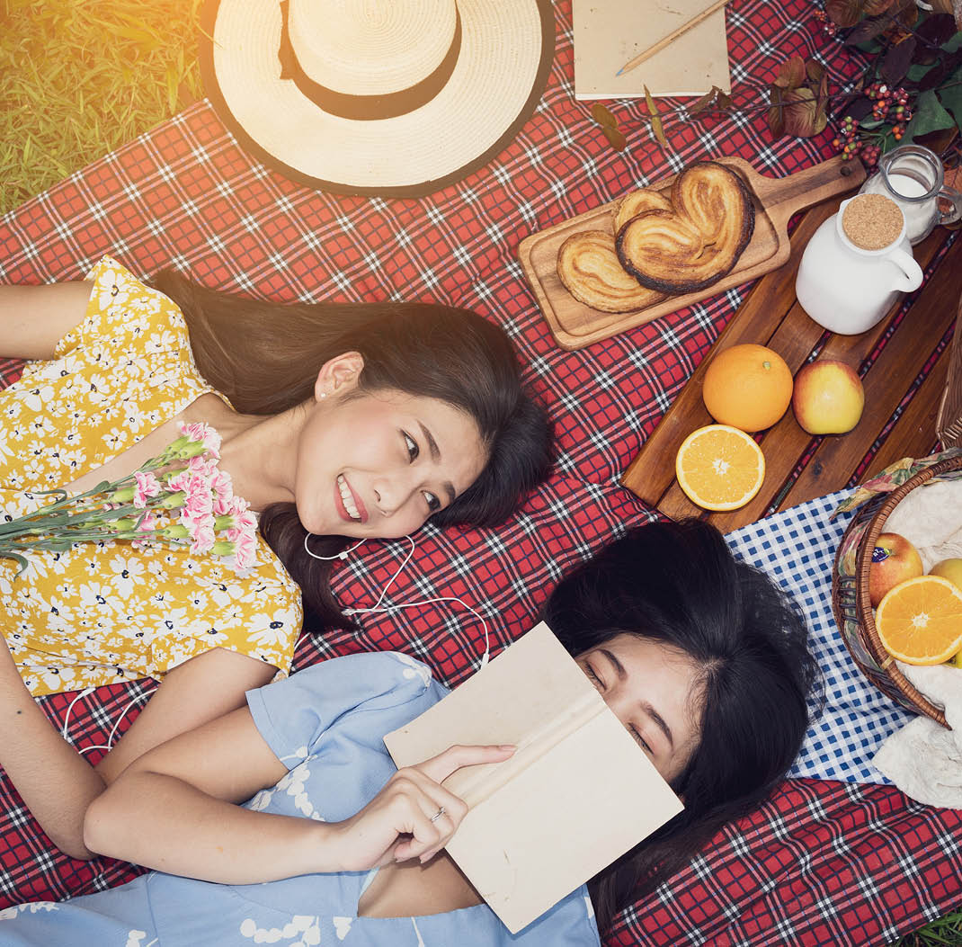 Couple of girls going picnic in a park, reading a book together and feeling friendly.
